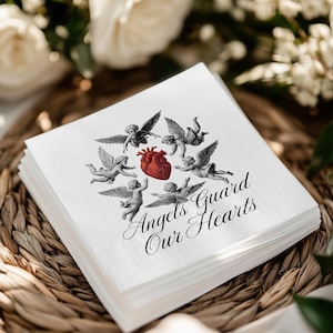 May include: A stack of white paper napkins with a design featuring cherubic angels surrounding a red heart. The text "Angels Guard Our Hearts" is printed in elegant script below the angels. The napkins are stacked on a woven basket.