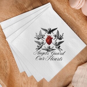 May include: White paper napkins with a design featuring cherubs surrounding a red heart. The text "Angels Guard Our Hearts" is printed below the cherubs. The napkins are stacked on a wooden surface.