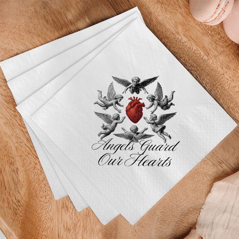 May include: White paper napkins with a design featuring cherubs surrounding a red heart. The text "Angels Guard Our Hearts" is printed below the cherubs. The napkins are stacked on a wooden surface.