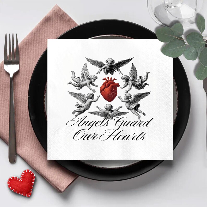 May include: A white square napkin with a design of cherubs surrounding a red heart, with the text "Angels Guard Our Hearts" in script. The napkin is on a black plate, with a pink napkin and a fork to the left.