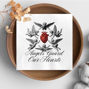 May include: White square napkin with a design featuring cherubic angels surrounding a red heart. The text "Angels Guard Our Hearts" is written in elegant script below the angels. The napkin is placed in a wooden bowl.
