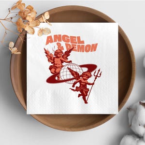 May include: White square napkin with the text "ANGEL & DEMON" in orange. The napkin features two cherubic figures, one with wings and the other holding a trident, with a globe and ring design in orange. The napkin is placed in a wooden bowl.