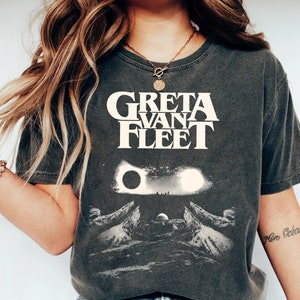 Può includere: Una maglietta grigia scura con una grafica bianca della band Greta Van Fleet. La grafica presenta il nome della band in lettere bianche in grassetto e un'immagine stilizzata di una catena montuosa con una luna crescente e due soli.