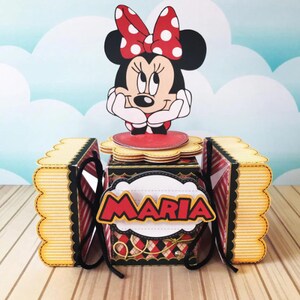 Peut inclure: Une bo&icirc;te en carton noire et rouge avec un personnage de Minnie Mouse sur le dessus. La bo&icirc;te a un bord festonn&eacute; et un motif ray&eacute; jaune et blanc. La bo&icirc;te est d&eacute;cor&eacute;e d'un ruban dor&eacute; et le nom "MARIA" est &eacute;crit sur le devant.