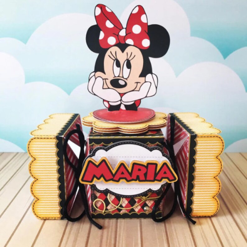 Peut inclure: Une bo&icirc;te en carton noire et rouge avec un personnage de Minnie Mouse sur le dessus. La bo&icirc;te a un bord festonn&eacute; et un motif ray&eacute; jaune et blanc. La bo&icirc;te est d&eacute;cor&eacute;e d'un ruban dor&eacute; et le nom "MARIA" est &eacute;crit sur le devant.
