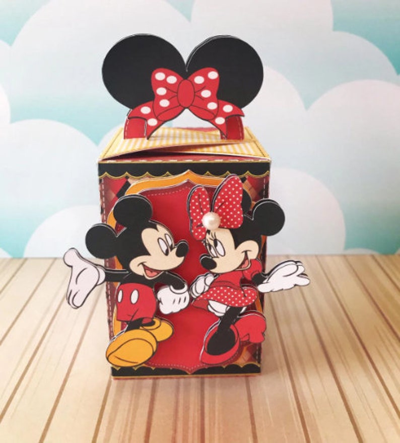 Peut inclure: Une bo&icirc;te en carton rouge et jaune avec une image 3D de Mickey Mouse et Minnie Mouse. Mickey Mouse porte un short rouge et des chaussures jaunes. Minnie Mouse porte une robe rouge &agrave; pois blancs et des chaussures rouges. La bo&icirc;te a un n&oelig;ud rouge &agrave; pois blancs sur le dessus.