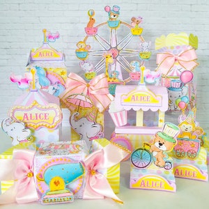 Könnte beinhalten: Ein Set an pink- und gelbfarbenen Partydekorationen mit Zirkus-Thema. Die Dekorationen beinhalten ein Riesenrad, einen Popcornstand, ein Karussell und eine Kanone. Die Dekorationen sind mit Bildern von Elefanten, Bären und anderen Zirkustieren verziert. Die Dekorationen sind mit dem Namen "Alice" beschriftet.
