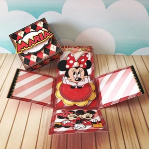 Peut inclure: Une bo&icirc;te &agrave; explosion rouge et noire avec une figurine Minnie Mouse pop-up. La bo&icirc;te est d&eacute;cor&eacute;e d'un motif &agrave; carreaux rouge et blanc et d'un motif &agrave; rayures rose et blanc. La bo&icirc;te est ouverte et la figurine pop-up est visible. La bo&icirc;te est &eacute;tiquet&eacute;e "Maria".