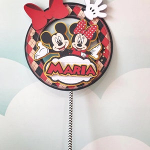 Peut inclure: Un topper de g&acirc;teau rouge, noir et blanc repr&eacute;sentant Mickey et Minnie Mouse. Le topper a un n&oelig;ud rouge, un gant blanc et un nuage jaune avec le nom "MARIA" &eacute;crit dessus.