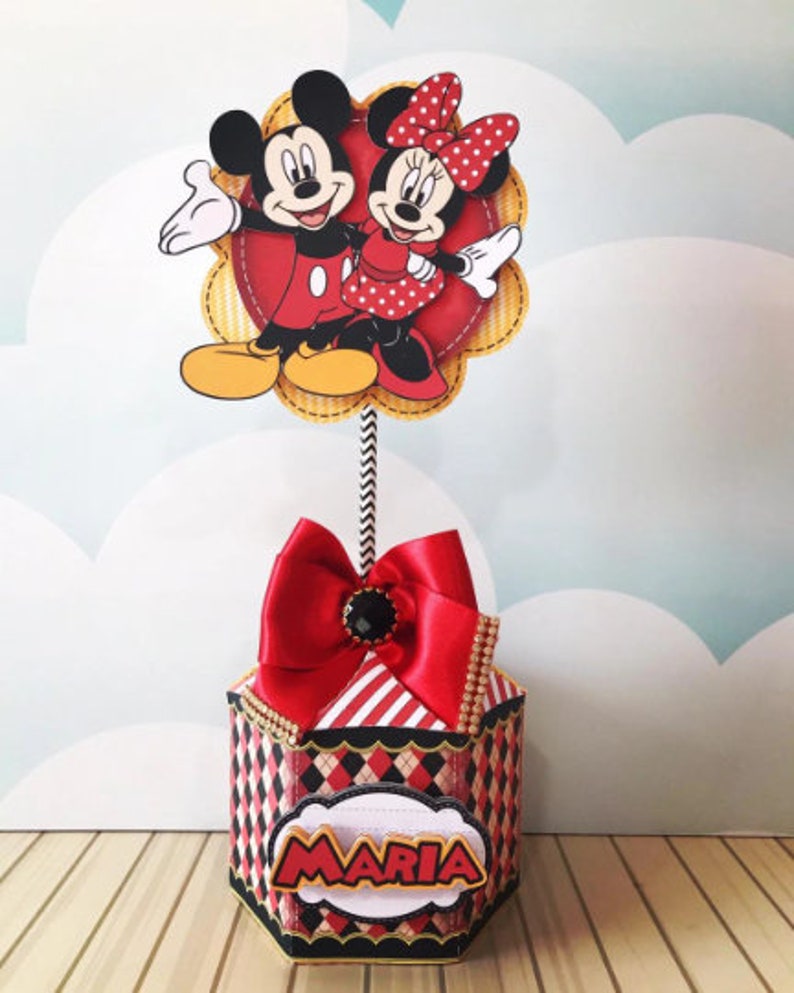 Peut inclure: Bo&icirc;te hexagonale rouge et noire avec un dessus ray&eacute; rouge et blanc et un n&oelig;ud rouge. La bo&icirc;te a une &eacute;tiquette blanche avec le nom "MARIA" en lettres dor&eacute;es. Un topper Mickey Mouse et Minnie Mouse est plac&eacute; sur le dessus de la bo&icirc;te.