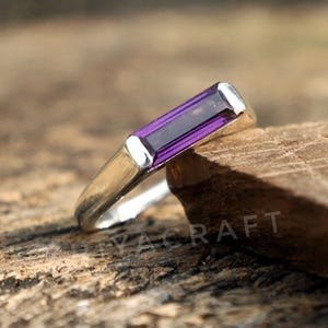 Könnte beinhalten: Ein silberner Ring mit einem rechteckigen, tiefvioletten Edelstein. Das Band des Rings ist glatt und poliert, und der Edelstein ist horizontal eingesetzt. Der Ring wird auf einem Holzstück präsentiert.