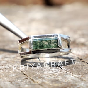 Puede incluir: Un anillo de plata con una piedra verde rectangular. El anillo tiene un diseño moderno con una forma geométrica. El texto "KRIYA CRAFT" es visible en la superficie. El anillo se muestra sobre una superficie de madera.