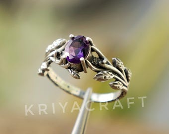 Peut inclure: Bague en argent ornée d'une pierre précieuse ovale violet foncé. La bague présente un motif inspiré de la nature avec des détails de feuilles et de vigne. Le texte "KRIYACRAFT" est visible.