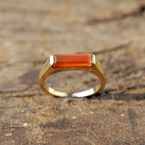 May include: A gold ring featuring a rectangular orange carnelian gemstone. The ring is displayed on a wooden surface.
