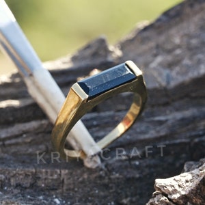 Könnte beinhalten: Ein goldener Ring mit einem schwarzen rechteckigen Edelstein. Der Ring liegt auf einer Holzoberfläche.