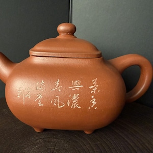 Könnte beinhalten: Eine terrakottafarbene Teekanne aus Ton mit matter Oberfläche, ausgestattet mit einem geschwungenen Griff, einem kurzen Ausguss und einem Deckel mit kleinem Knauf. Der Körper der Kanne ist vorne mit weißen chinesischen Schriftzeichen graviert und steht auf vier kleinen Füßen.