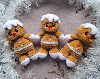 May include: Three crocheted gingerbread men with white icing details, red noses, and button accents. They are arranged on a light brown background, surrounded by a white, snowflake-adorned wreath. A festive, handmade holiday decoration.