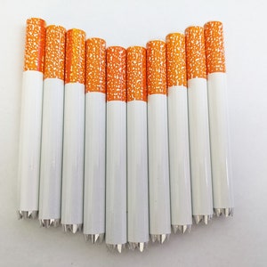 May include: A collection of white pencils with orange and white speckled tips, arranged in a curved line. The pencils have silver-colored, pointed ends. The pencils are set against a plain white background.