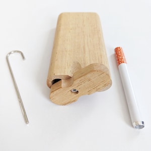 May include: A wooden cigarette case with a matching wooden lid, a metal poker, and a white cigarette with an orange filter. The case is light brown and has a carved design on the front. The cigarette is white with a silver tip.