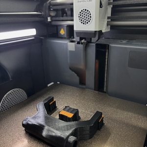 May include: A 3D printer with the text "Bambu Lab" on the front. The printer is printing a black and orange object. The object is on a textured surface.