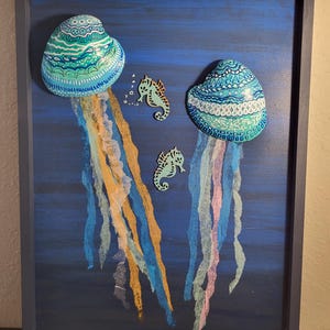May include: A framed artwork featuring two stylized jellyfish with patterned caps in shades of blue and teal. The jellyfish have flowing, colorful tentacles in yellow, blue, and pink. Two seahorse cutouts are also included in the artwork, set against a dark blue background.