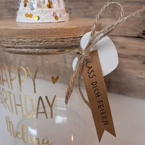 Op de afbeelding: Een heldere glazen pot met gouden letters die "HAPPY BIRTHDAY Melina" leest en een klein gouden hartje. De pot heeft een houten deksel, een witte kaarsenhouder met gouden en zilveren confetti en een label met "LASS DICH FEIERN".
