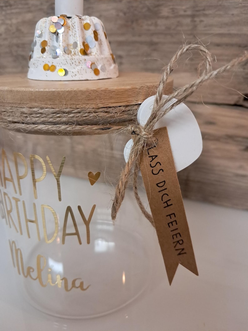 Op de afbeelding: Een heldere glazen pot met gouden letters die "HAPPY BIRTHDAY Melina" leest en een klein gouden hartje. De pot heeft een houten deksel, een witte kaarsenhouder met gouden en zilveren confetti en een label met "LASS DICH FEIERN".