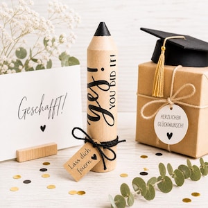 Könnte beinhalten: Ein Geschenkset zum Thema Abschluss mit einem großen stiftförmigen Behälter mit der Aufschrift "Yes! You Did It!", einer kleinen Abschlusskappe und einer Geschenkbox. Eine Karte mit "Geschafft!" ist ebenfalls vorhanden.
