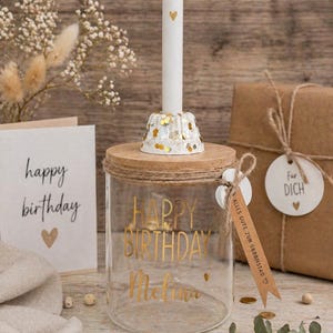 Op de afbeelding: Een glazen pot met houten deksel en gouden tekst "HAPPY BIRTHDAY Melina". Een witte kaars met een gouden hart in een decoratieve houder. Een verjaardagskaart en een cadeau zijn ook aanwezig, wat een feestelijke sfeer cre&euml;ert.