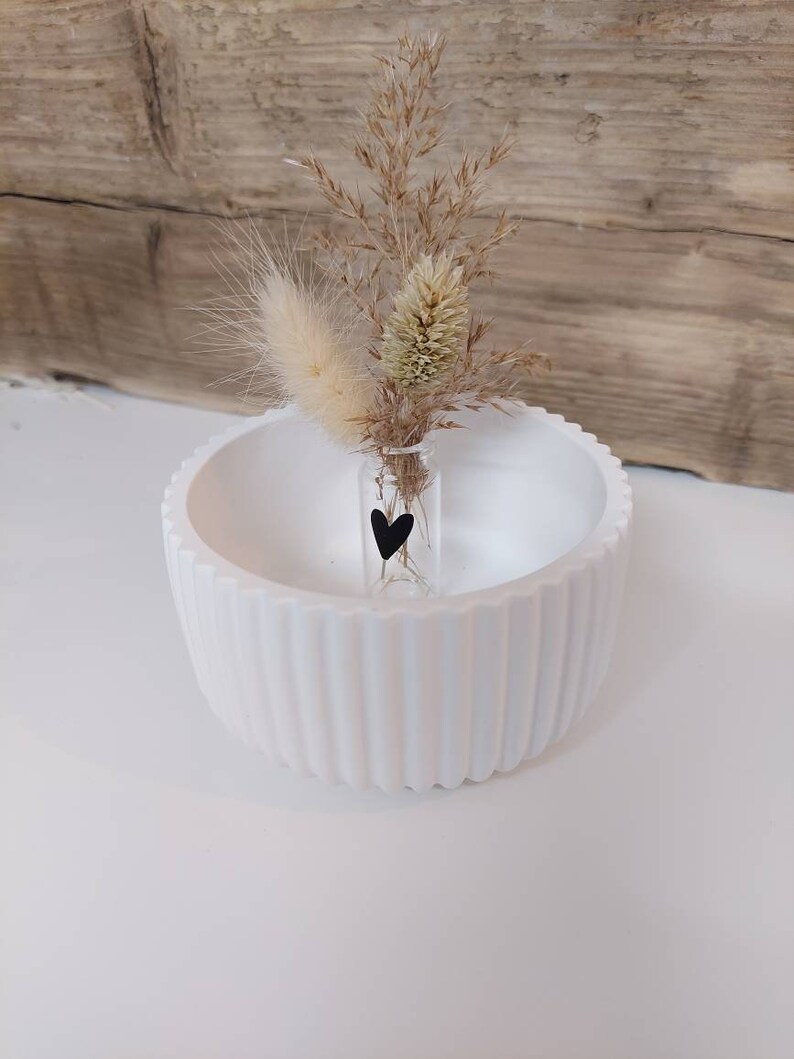 Puede incluir: Un cuenco de cer&aacute;mica blanco con un dise&ntilde;o acanalado, que contiene un peque&ntilde;o jarr&oacute;n de vidrio con hierba de la pampa seca y una pegatina de coraz&oacute;n negro.