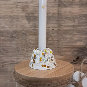 Op de afbeelding: Een witte kaars met een gouden hartontwerp, geplaatst in een witte kandelaar versierd met gouden en zilveren confetti. De houder staat op een houten basis, onderdeel van een decoratieve pot.