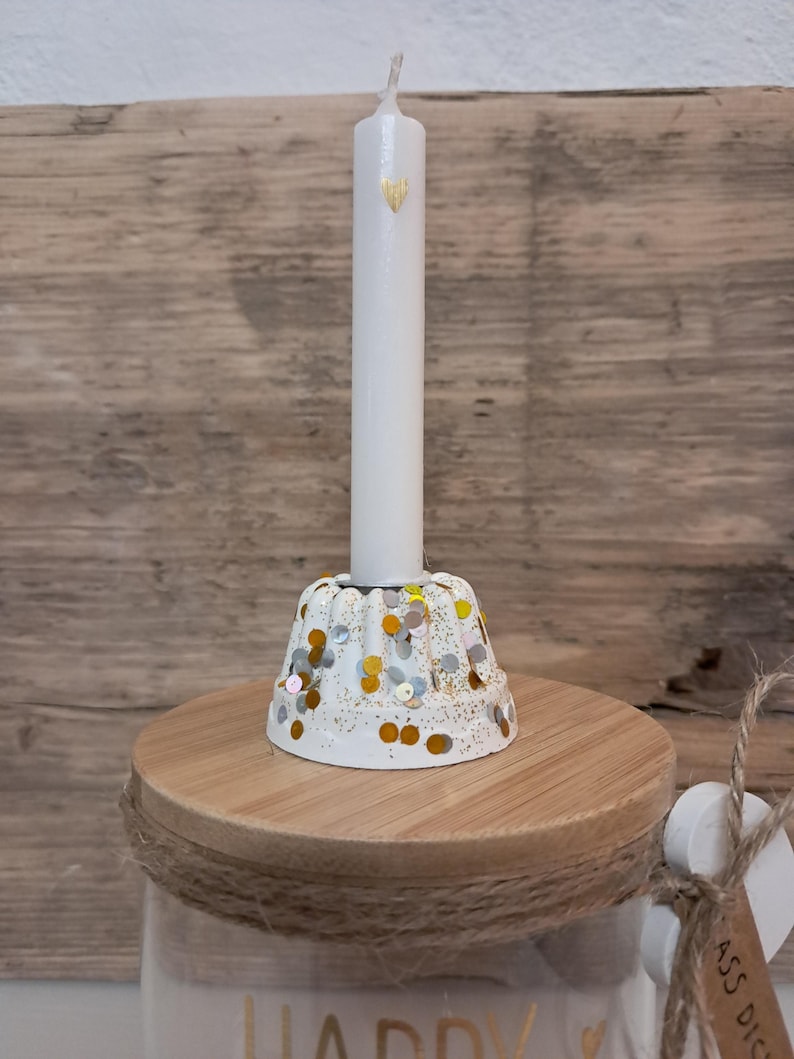 Op de afbeelding: Een witte kaars met een gouden hartontwerp, geplaatst in een witte kandelaar versierd met gouden en zilveren confetti. De houder staat op een houten basis, onderdeel van een decoratieve pot.