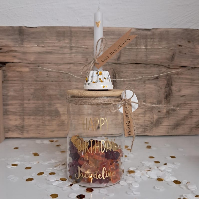 Op de afbeelding: Een glazen pot gevuld met kleurrijke gummy snoepjes, een houten deksel en een kaars. De pot heeft gouden letters met de tekst "Happy Birthday Jacqueline". Een klein label leest "F&uuml;r Dich". Een kandelaar is versierd met goud en zilver glitter.