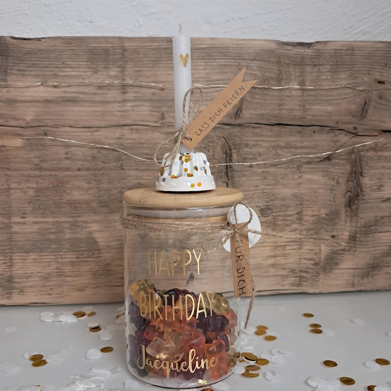 Op de afbeelding: Een glazen pot gevuld met gummy snoepjes, een houten deksel en een witte kaars met een gouden hart. De pot is versierd met gouden letters met de tekst "Happy Birthday Jacqueline". Een klein label leest "Voor jou".