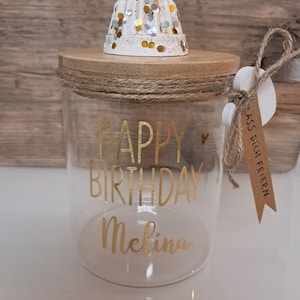 Op de afbeelding: Een heldere glazen pot met een houten deksel en touwdetails. De pot heeft gouden letters met de tekst "HAPPY BIRTHDAY Melina". Bovenop staat een witte kandelaar met gouden en zilveren confetti. Een klein label is bevestigd.
