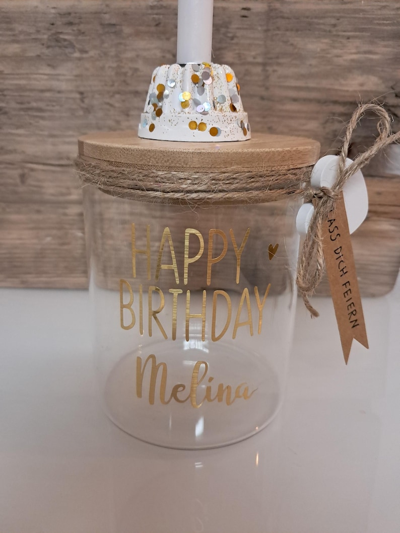 Op de afbeelding: Een heldere glazen pot met een houten deksel en touwdetails. De pot heeft gouden letters met de tekst "HAPPY BIRTHDAY Melina". Bovenop staat een witte kandelaar met gouden en zilveren confetti. Een klein label is bevestigd.