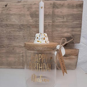 Op de afbeelding: Een heldere glazen pot met een houten deksel, met gouden letters die "HAPPY BIRTHDAY Melina" lezen. Een witte kaars met een hartontwerp staat op een decoratieve houder met gouden en zilveren confetti. Een klein label is bevestigd.