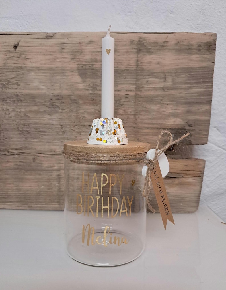 Op de afbeelding: Een heldere glazen pot met een houten deksel, met gouden letters die "HAPPY BIRTHDAY Melina" lezen. Een witte kaars met een hartontwerp staat op een decoratieve houder met gouden en zilveren confetti. Een klein label is bevestigd.