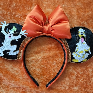 Può includere: Un paio di orecchie di Topolino nere e dorate con un grande fiocco arancione. Le orecchie mostrano un Goofy e un Pluto mummificati.