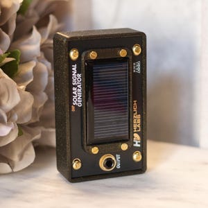 May include: A black rectangular solar signal generator with a small solar panel and gold-coloured screws. The text "OHP SOLAR SIGNAL GENERATOR" and "Herzlich Labs" are printed on the device. The device has an output jack.