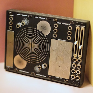 Könnte beinhalten: Ein schwarzes elektronisches Musik-Synthesizer-Modul mit goldenen Akzenten. Das Modul hat zwei Knöpfe, eine Reihe von Buchsen und ein grafisches Design mit dem Text "MMI HELIOS", "MMI MATRIX" und "HERZLICH".