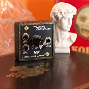 May include: A black and gold OHP Passive Crossfader by Herzlich Labs. The device has input and output jacks, a central fade knob, and gold-colored accents. The device is sitting on a decorative surface.