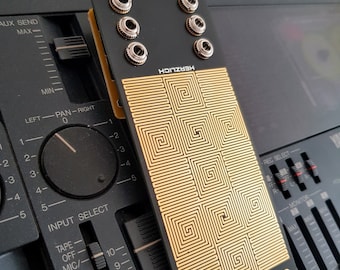 May include: A black and gold electronic music module with the text "MMI MATRIX" and "HERZLICH" printed on it. The module has a grid of small squares with a spiral pattern.