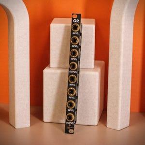 May include: A black electronic module with gold-colored jacks and the text "OR" at the top. The module has multiple "INPUT" and "OUTPUT" labels. It is positioned vertically against a backdrop of white cubes and an orange background.