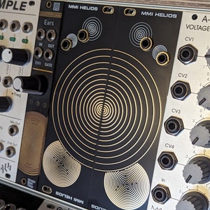 Könnte beinhalten: Ein schwarzes und goldenes MMI HELIOS Eurorack-Modul mit einem kreisförmigen Muster und mehreren Knöpfen und Buchsen. Das Modul trägt den Text "MMI HELIOS".