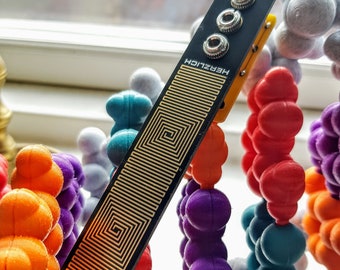 May include: A black circuit board with gold accents and the text "HERZLICH" and "MMI". The board is resting on a colorful plastic toy structure.