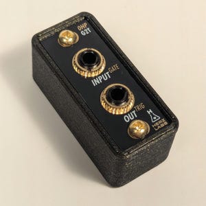 Puede incluir: Caja rectangular negra con tres botones dorados y dos conectores de audio negros. La caja está etiquetada con "OHP G2T", "INPUT GATE", "OUT TRIG" y "HERZ LABS".