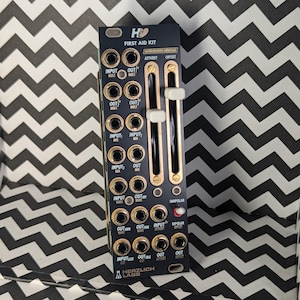 Könnte beinhalten: Ein schwarzes und goldenes Eurorack-Modul mit dem Text "He FIRST AID KIT" und "INTERVENTION MEDICALE" darauf gedruckt. Das Modul hat mehrere Knöpfe, Buchsen und Schalter.