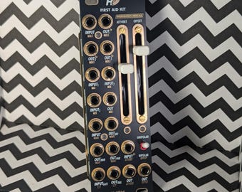 May include: A black and gold Eurorack module with the text "He FIRST AID KIT" and "INTERVENTION MEDICALE" printed on it. The module has multiple knobs, jacks, and switches.