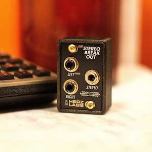 May include: A small, black audio interface with gold-coloured jacks and the text "STEREO BREAK OUT." It features inputs marked "LEFT mono," "RIGHT," and "STEREO." The brand name "HERZ LABS" is also visible.